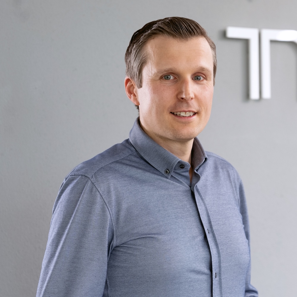 Dr. André Kortmann in a light blue button-up shirt standing in front of a gray wall.