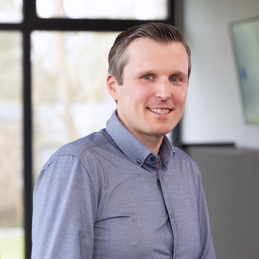 Dr. André Kortmann in a light blue button-up shirt is standing in a bright, modern office environment with large windows in the background.