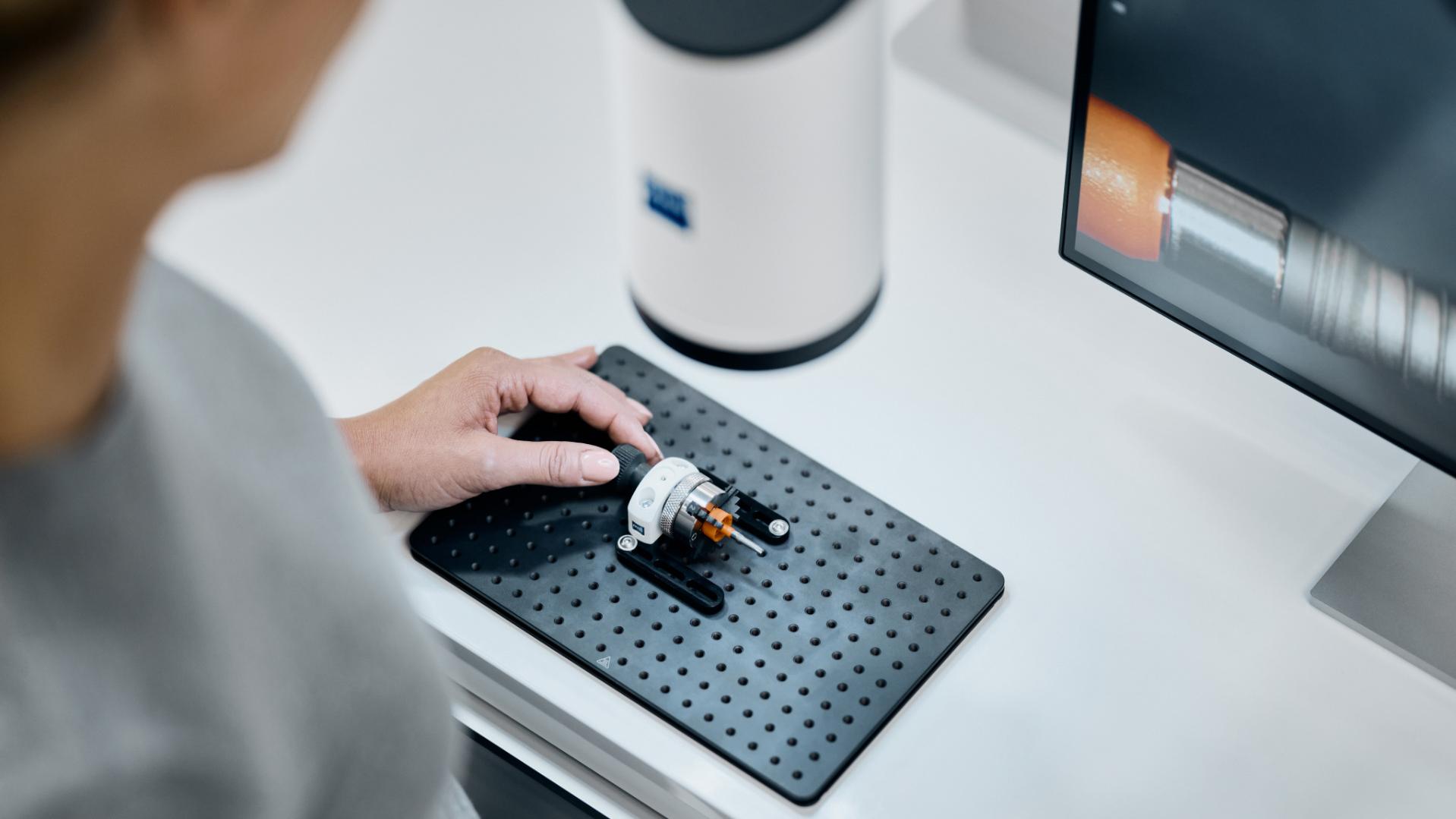 A person operates a ZEISS Smartzoom 100 to inspect a small mechanical component on a perforated work surface next to a monitor displaying analysis data.