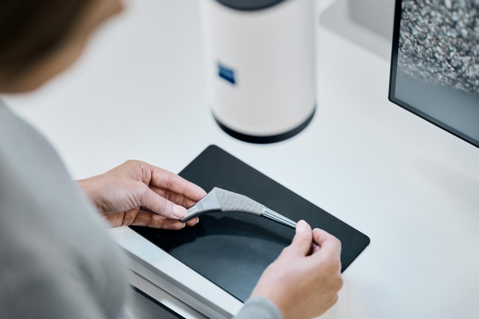 A person holding a 3D-printed part on a black surface next to a ZEISS device and a monitor displaying a magnified material structure.