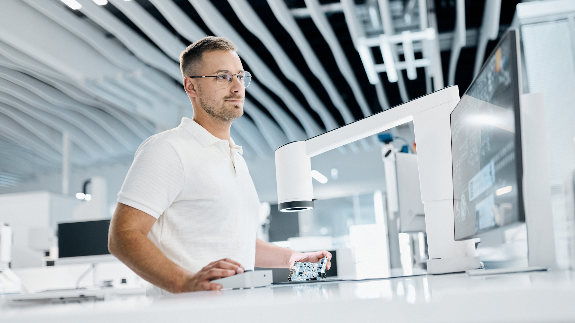A man in a white polo shirt examines a circuit board under a digital microscope ZEISS Smartzoom 100 at a ZEISS laboratory workstation.