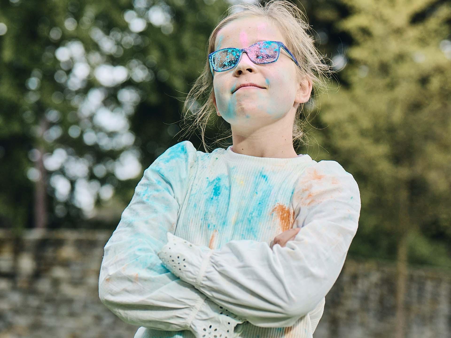 A girl with crossed arms and diry glasses from playing with colored poweder looks boldly and smirks.