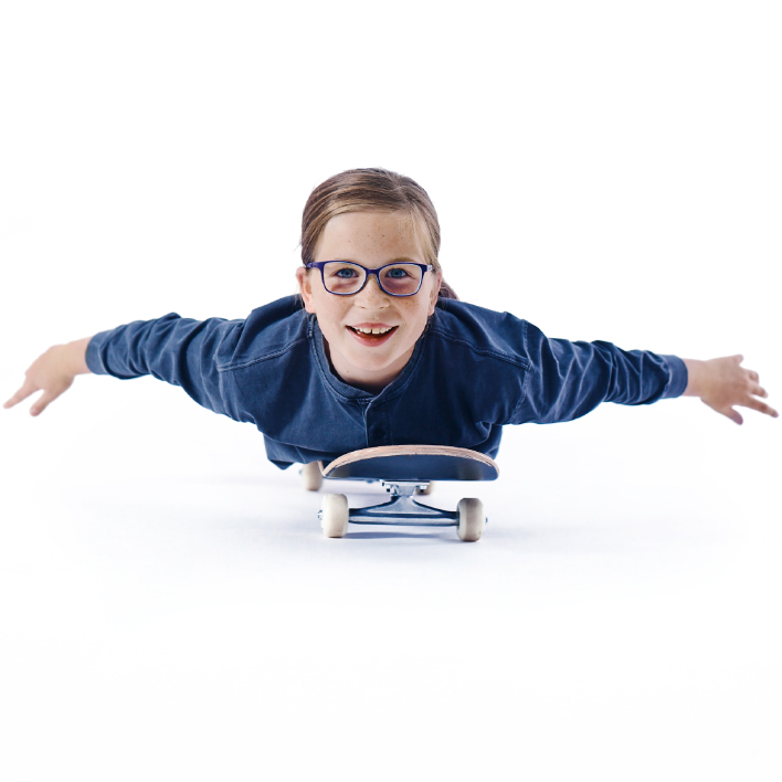 A young girl wearing glasses lies flat on her skateboard.